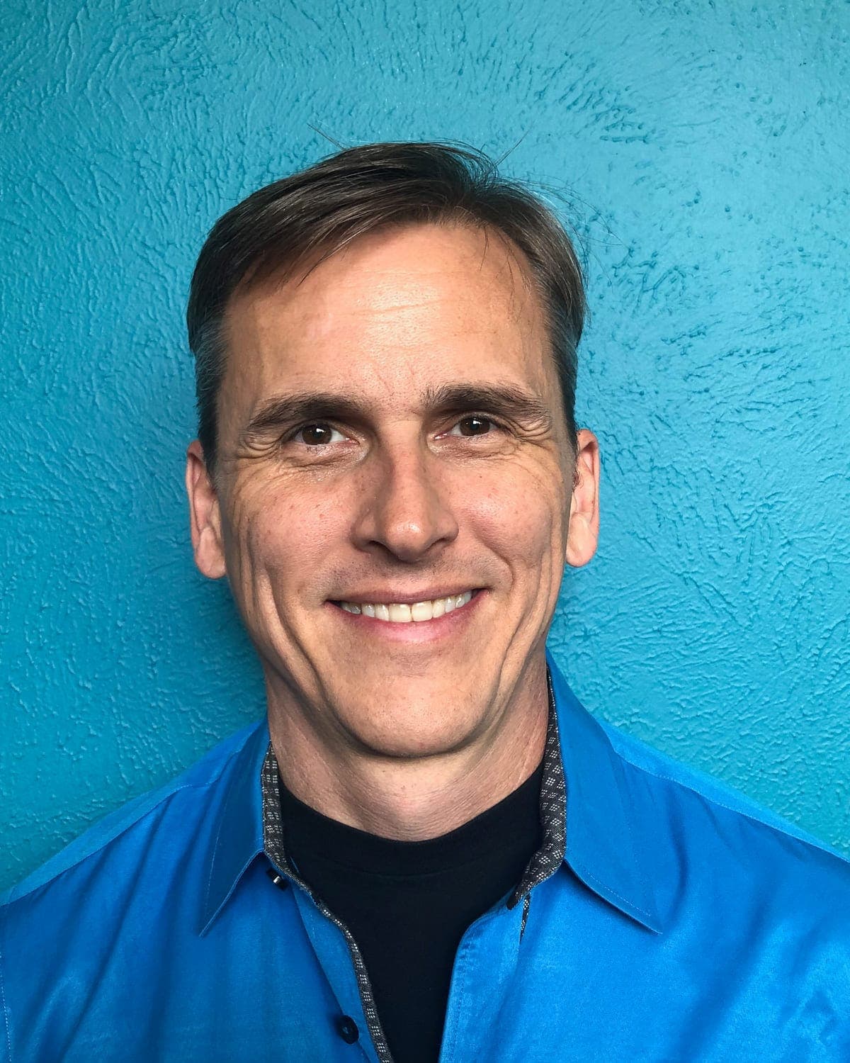 Bruce Ultra, smiling, in a blue shirt against a teal wall
